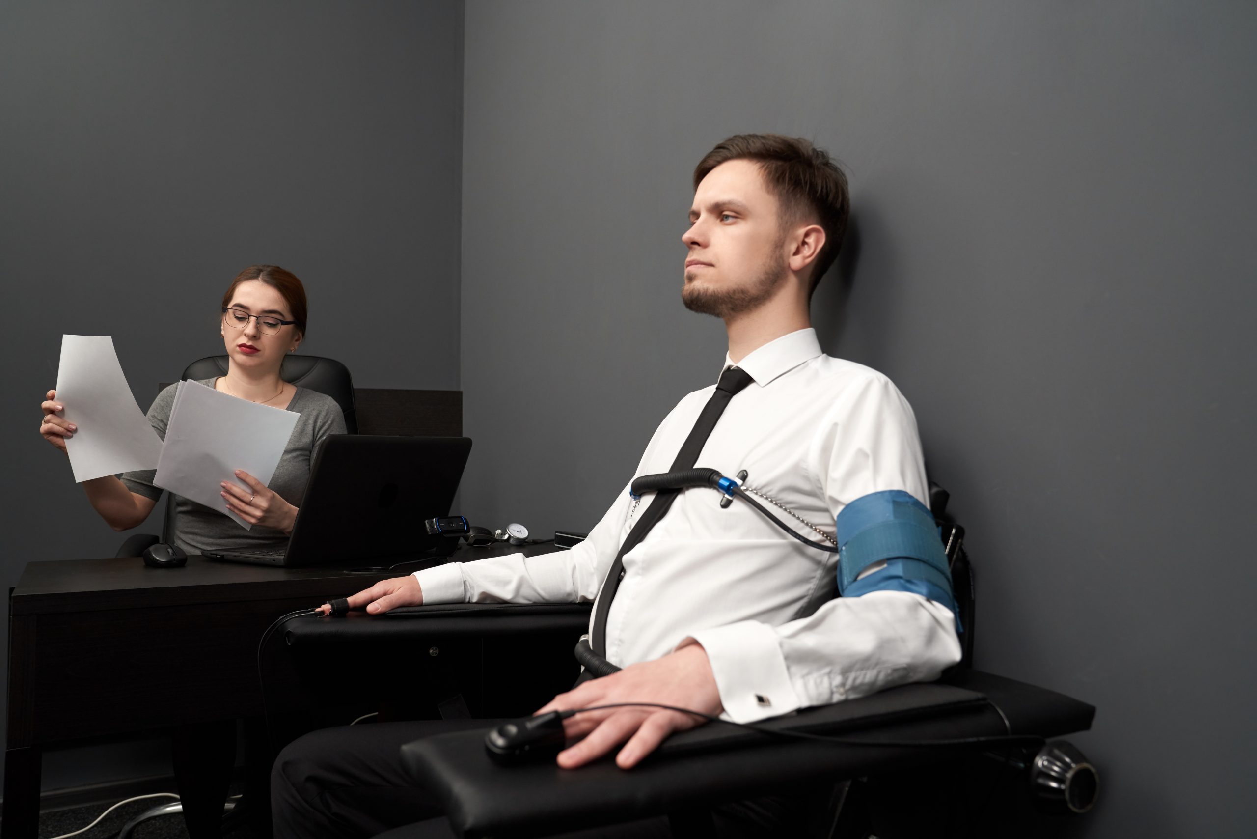 Handsome man sitting in chair in grey room and testing with polygraph. To hands and body attached sensors. Young woman in glasses sitting at table with computer polygraph, looking at papers.